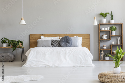 Plant on pouf in bright bedroom interior with pillows on bed with wooden headboard. Real photo