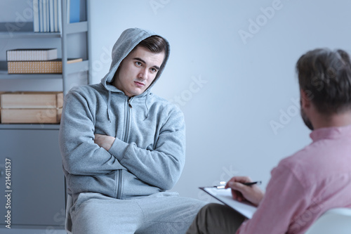 An individual therapeutic session between a psychologist and a withdrawn troubled teenage boy sitting in a closed position with his arms crossed across his chest.