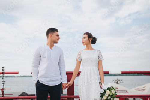 Beautiful newlyweds stand on the waterfront on the background of the water and the city. The stylish bridegroom holds the bride's hand. Wedding.