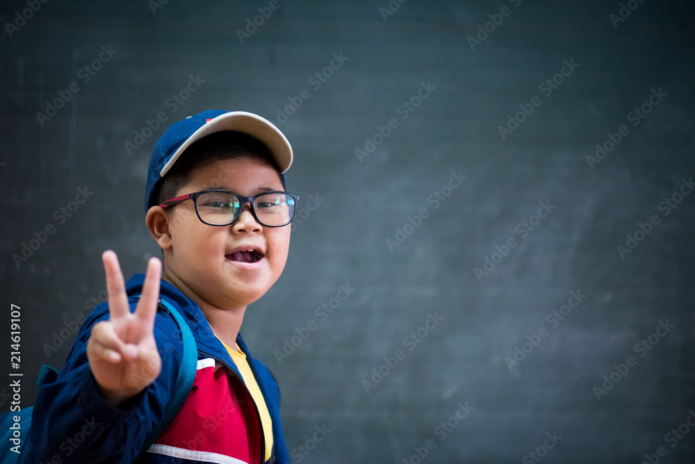Happy smiling asian boy in glasses showing two fingers as victory sign is going to school for the first time. Child with school bag and book on blackboard background. Back to school concept.