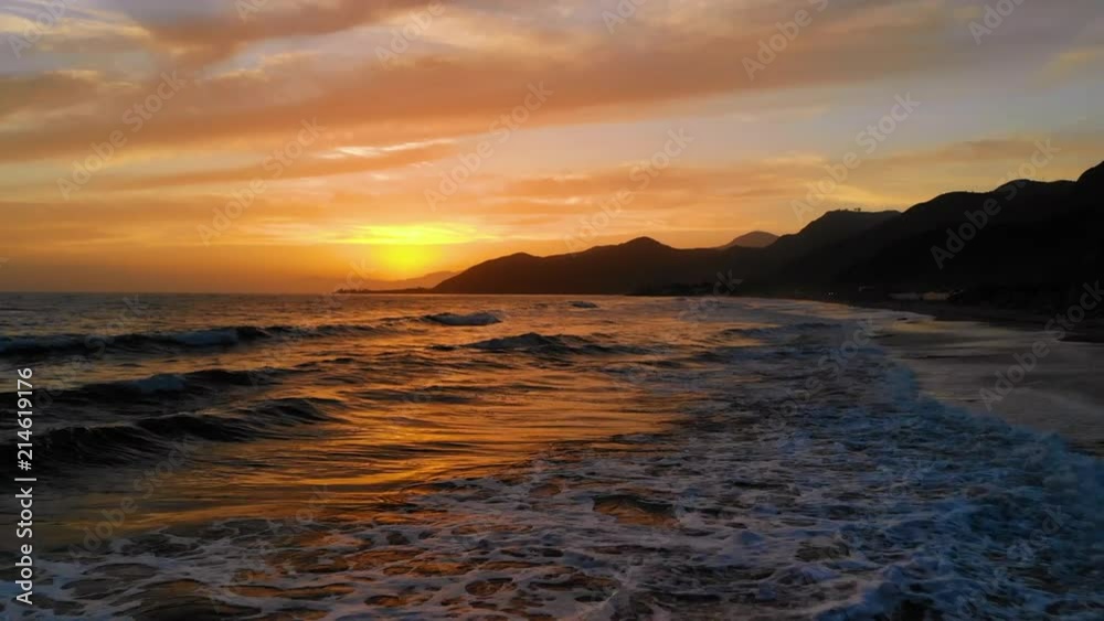 AERIAL drone shot off of the California coast at sunset with bright orange sky and ocean waves underneath on the beach