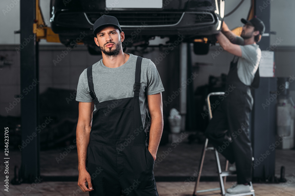 auto mechanic posing in overalls, while colleague working in workshop behind auto mechanic posing in overalls, while colleague working in workshop behind