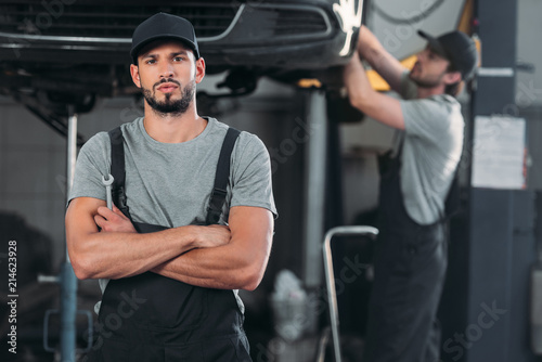 serious mechanic with crossed arms holding wrench, while colleague working in workshop behind