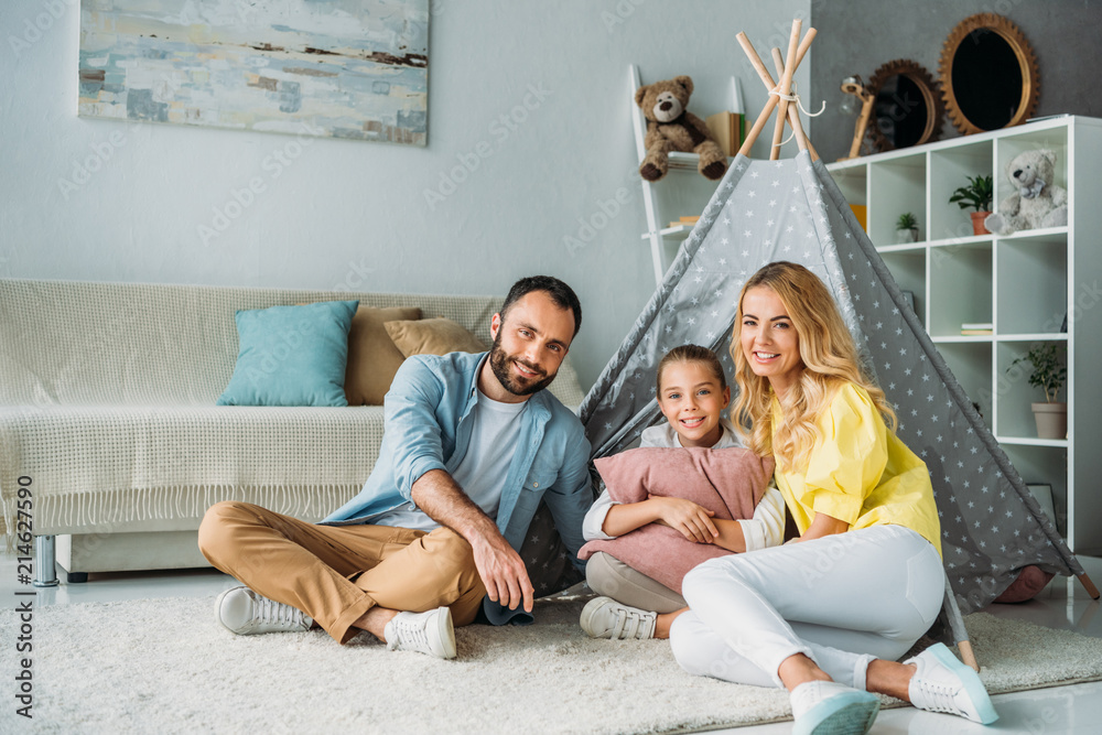 happy young family sitting on floor with teepee and looking at camera happy young family sitting on floor with teepee and looking at camera