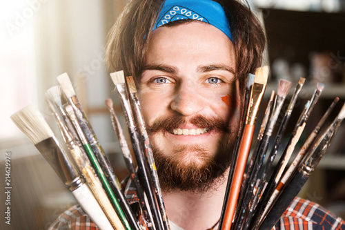 Dark-haired caucasian bearded hipster male artist in blue bandana hoilding many brushes in light studio photo