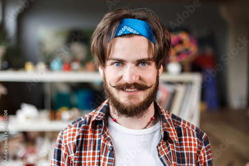 Portrait of handsome dark-haired bearded hipster male artist in blue bandana in his contemporary studio with many paintings photo