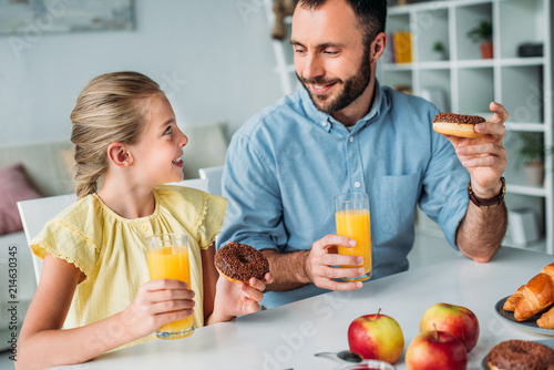 happy father and daughter eating doughnuts with orange juice at home