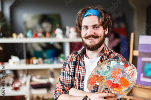 Portrait of handsome dark-haired bearded hipster male artist in blue bandana holding palette in his contemporary studio with many paintings photo