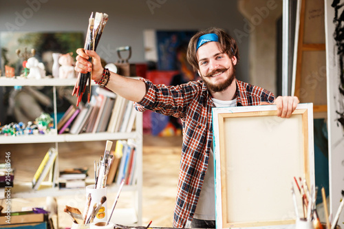 Handsome dark-haired bearded hipster male artist in blue bandana holding brush and smiling in his contemporary studio with many paintings, photo