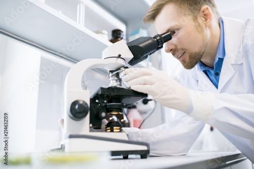 Side view portrait of smiling scientist looking in microscope while doing research in medical laboratory