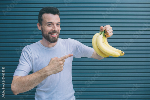 Happy guy is hlding bananas in one hand and pointing on them with another one. He is looking on camera and smiling. Isolated on striped and blue background. photo