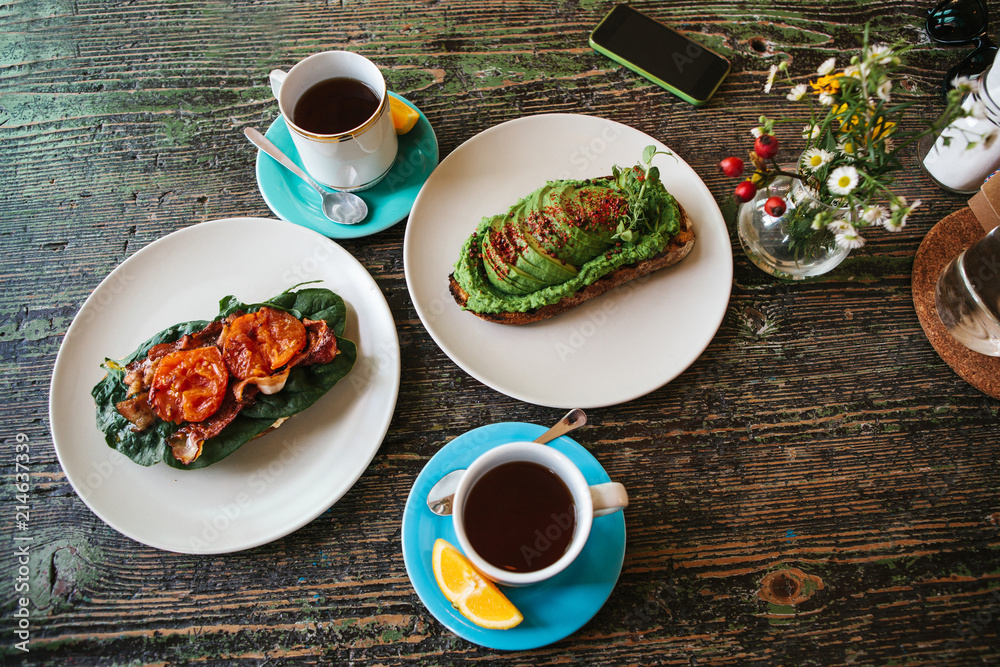 Sandwich with avocado and another sandwich with bacon and tomato next to two cups of tea on a wooden table Sandwich with avocado and another sandwich with bacon and tomato next to two cups of tea on a wooden table