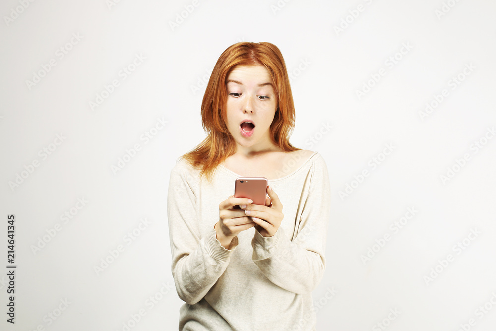 Surprised natural redhead woman with open mouth wearing white oversized loose shirt staring at cell phone screen. amazed hipster ginger female w/ smartphone. Isolated background, copy space, portrait. Surprised natural redhead woman with open mouth wearing white oversized loose shirt staring at cell phone screen. amazed hipster ginger female w/ smartphone. Isolated background, copy space, portrait.
