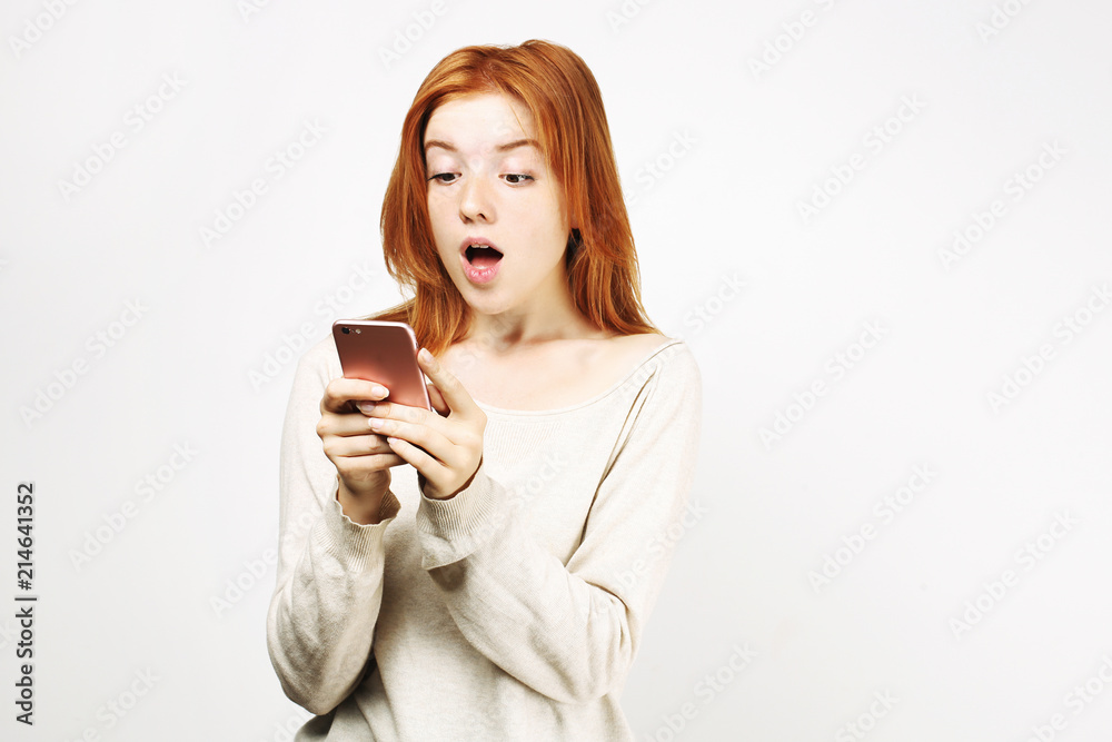 Surprised natural redhead woman with open mouth wearing white oversized loose shirt staring at cell phone screen. amazed hipster ginger female w/ smartphone. Isolated background, copy space, portrait. Surprised natural redhead woman with open mouth wearing white oversized loose shirt staring at cell phone screen. amazed hipster ginger female w/ smartphone. Isolated background, copy space, portrait.