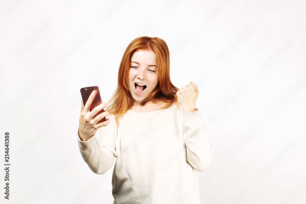 Lucky beautiful young happy woman with natural long red hair celebrating her win, screaming and laughing, looking at her smartphone screen. Attractive female in winning pose. Copy space background. Lucky beautiful young happy woman with natural long red hair celebrating her win, screaming and laughing, looking at her smartphone screen. Attractive female in winning pose. Copy space background.