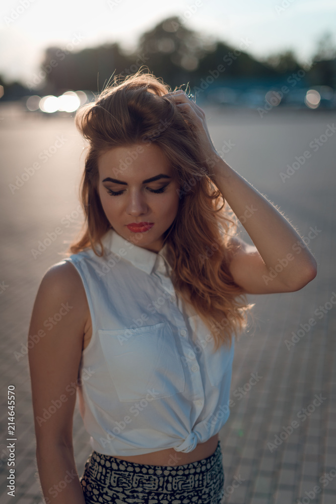 Close up portrait of beautiful blonde woman with curly hair, make up in white blouse top posing on the street on sunset