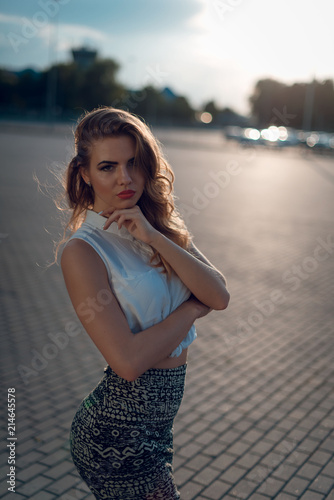 Portrait of attracive blonde woman with curly hair, beautiful make up, red lips, in white blouse top posing on the street on sunset. Close up © WellStock