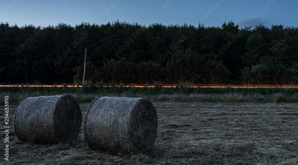 Car light streaks passing a road near a Field of hay bales at night