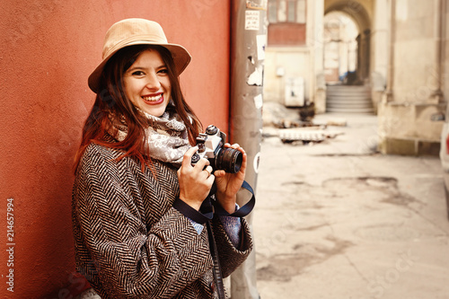 happy and stylish hipster woman holding film photo camera and smiling on background of old red wall building in european city