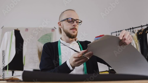 Close-up of a young beared designer or tailor in eyeglasses who sitting at a workplase at the studio and carving out with scissors a sketches for sewing a new collection of clothes. A stylish bearded photo