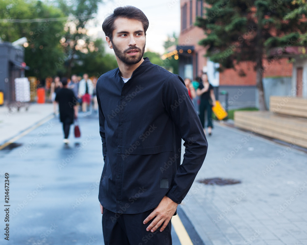 Youth Men's Fashion. A young male hippy with a beard posing on a summer evening outdoors in a fashionable outfit against a gray concrete wall in an art space. Black shirt and black trousers. Cover. Youth Men's Fashion. A young male hippy with a beard posing on a summer evening outdoors in a fashionable outfit against a gray concrete wall in an art space. Black shirt and black trousers. Cover.