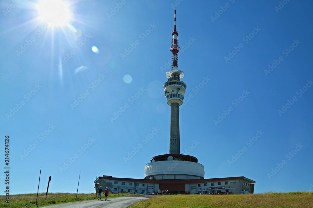 Fototapeta premium Red and white telecommunication and lookout tower Praded, Czech Republic, Europe standing in Jeseniky mountains, backlit with sun, tourists hiking on road, sunny summer day clear blue sky, green trees
