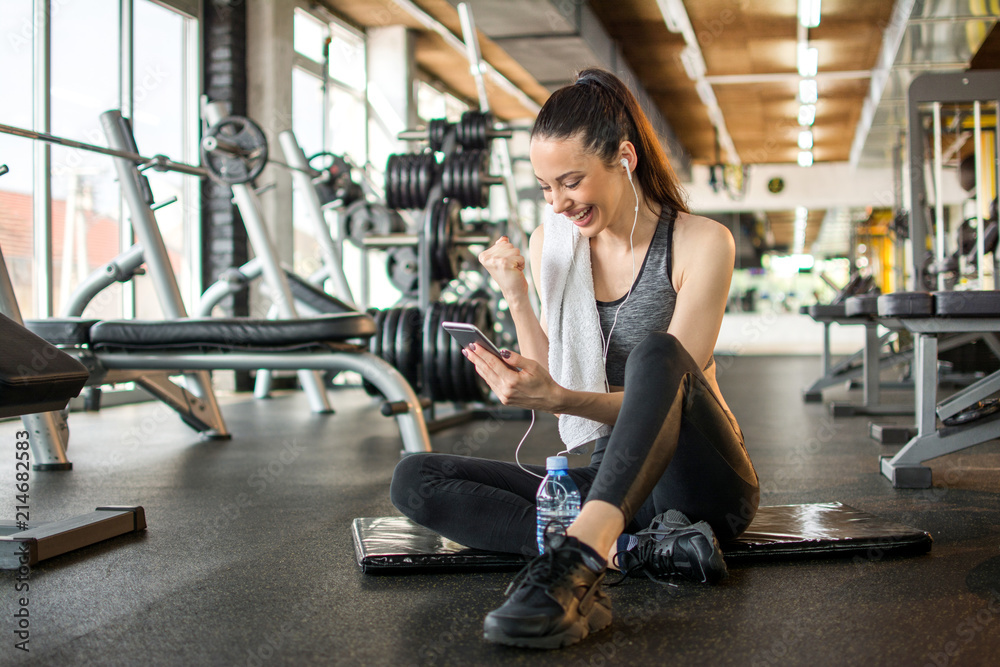Cheerful young woman reading good news on mobile phone and celebrating success during workout break in gym Cheerful young woman reading good news on mobile phone and celebrating success during workout break in gym