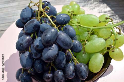 Two brushes of grapes on a platter. Blue and green grapes. The dish is on a round pink table. Open summer terrace. A beautiful sunny day.