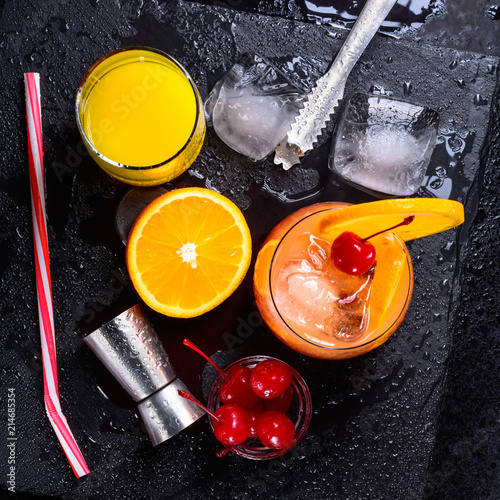 Tequila Sunrise cocktail, orange, ice cubes, maraschino cherries, ice tongs, jigger and straw on a black wet slate tray. Cocktail Set. Top view. photo