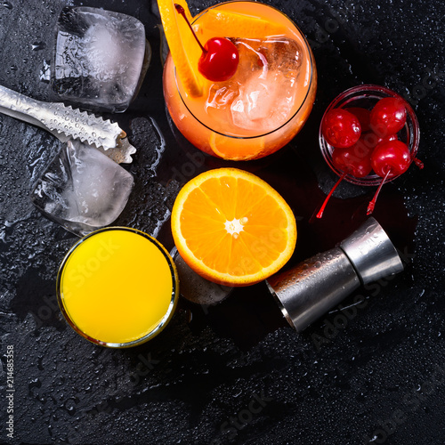 Tequila Sunrise cocktail, orange, ice cubes, maraschino cherries, ice tongs and jigger on a wet black slate tray. Cocktail Set. Top view. photo