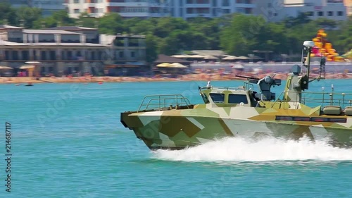 Close-up military landing speed boat with weapons quickly floats on blue surface of sea leaving traces of foam on background of shore and buildings