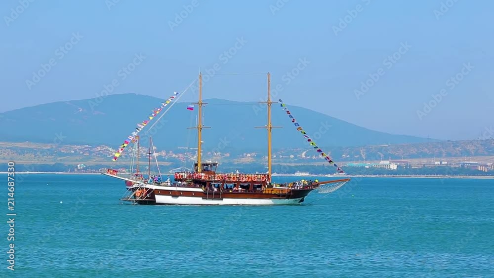 Beautiful huge wooden ship in pirate style drifts on blue surface of sea against sea coast and mountains under the boundless blue sky Beautiful huge wooden ship in pirate style drifts on blue surface of sea against sea coast and mountains under the boundless blue sky