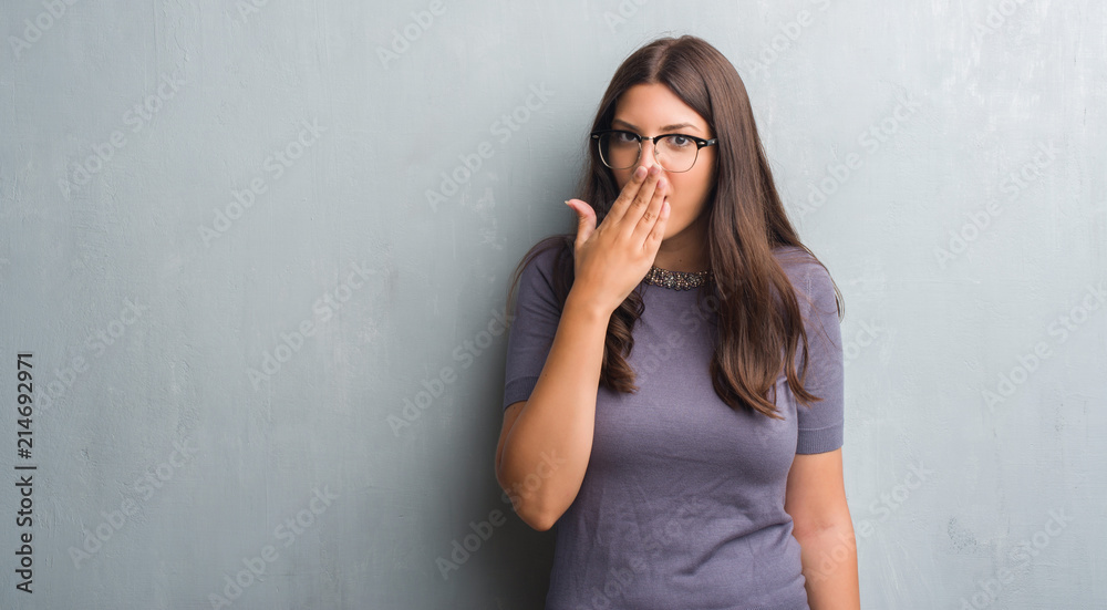 Young brunette woman over grunge grey wall wearing glasses cover mouth with hand shocked with shame for mistake, expression of fear, scared in silence, secret concept Young brunette woman over grunge grey wall wearing glasses cover mouth with hand shocked with shame for mistake, expression of fear, scared in silence, secret concept