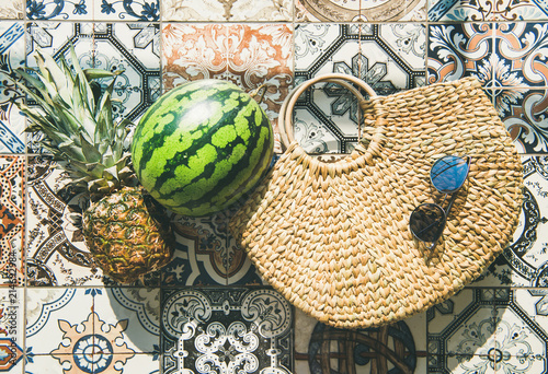 Summer lifestyle background. Flat-lay of summer fruit pineapple and watermelon, straw bag and sunglasses over colorful moroccan tile floor, top view, horizontal composition