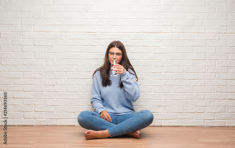 Young brunette woman sitting on the floor drinking glass of water with a confident expression on smart face thinking serious Young brunette woman sitting on the floor drinking glass of water with a confident expression on smart face thinking serious