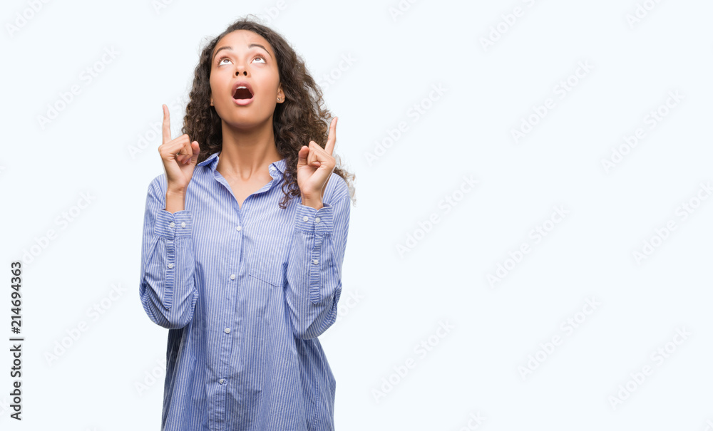 Young hispanic business woman amazed and surprised looking up and pointing with fingers and raised arms. Young hispanic business woman amazed and surprised looking up and pointing with fingers and raised arms.