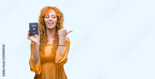 Young redhead woman holding passport of Germany pointing and showing with thumb up to the side with happy face smiling