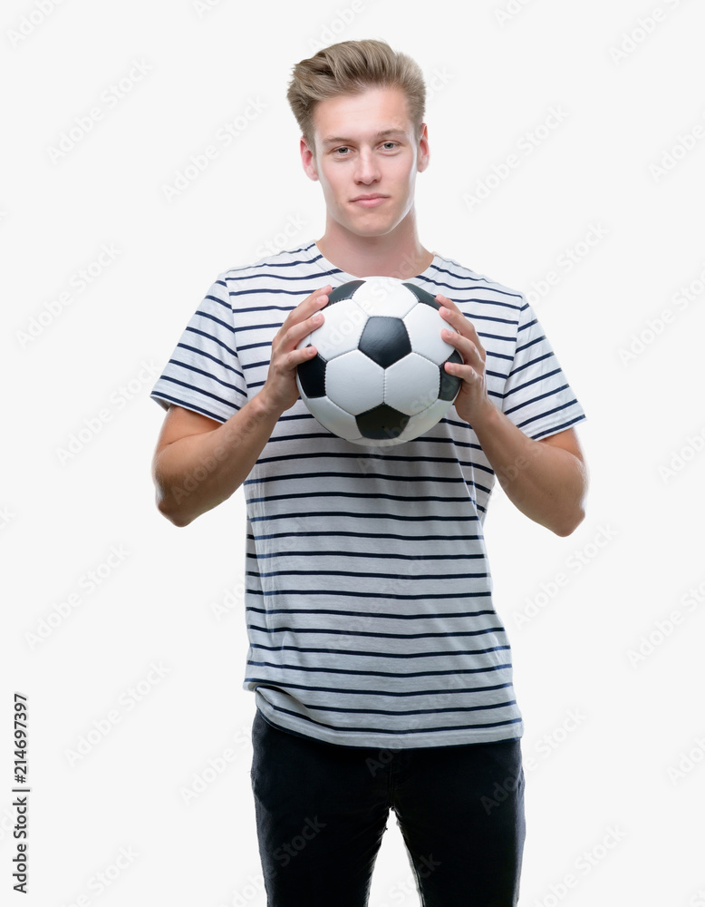 Young handsome blond man holding soccer ball with a confident expression on smart face thinking serious