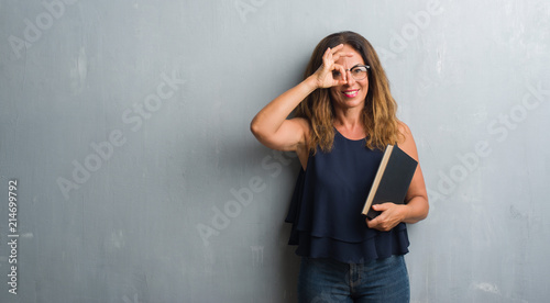 Middle age hispanic woman standing over grey grunge wall holding a book with happy face smiling doing ok sign with hand on eye looking through fingers