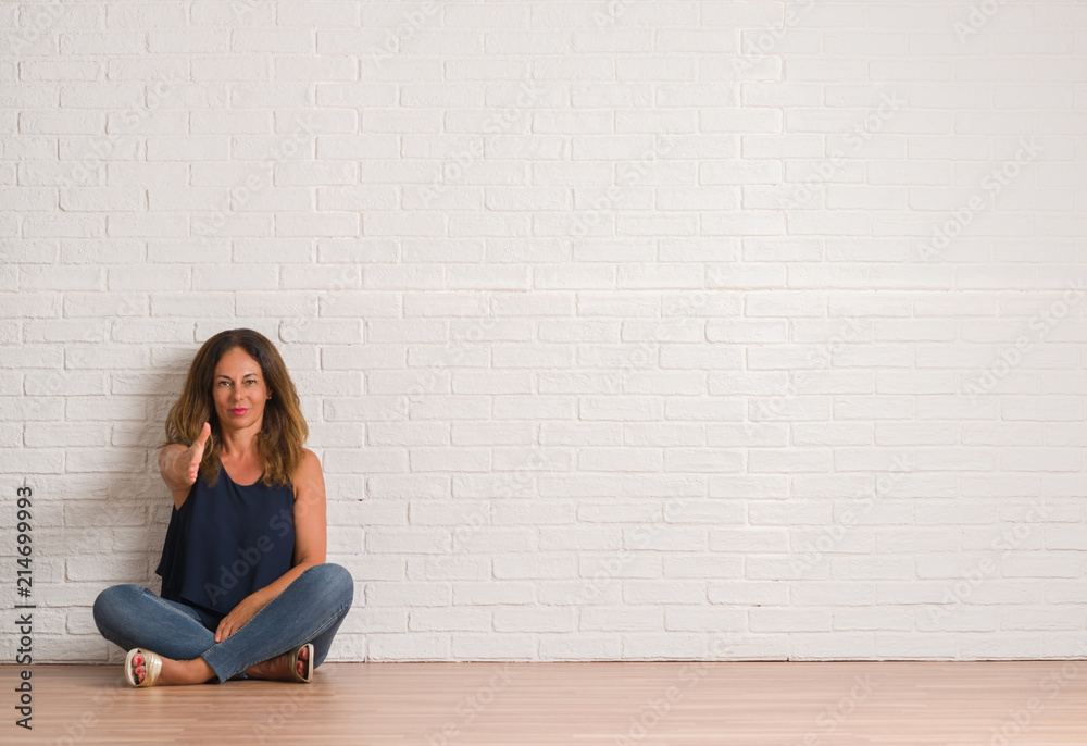 Middle age hispanic woman sitting on the floor over white brick wall smiling friendly offering handshake as greeting and welcoming. Successful business. Middle age hispanic woman sitting on the floor over white brick wall smiling friendly offering handshake as greeting and welcoming. Successful business.