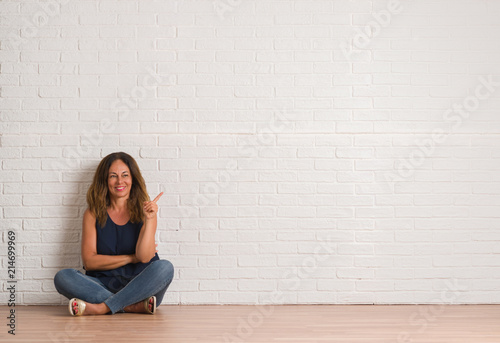 Middle age hispanic woman sitting on the floor over white brick wall with a big smile on face, pointing with hand and finger to the side looking at the camera. photo