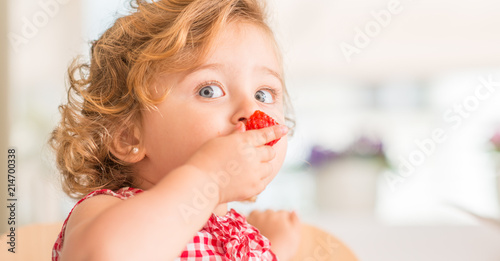 Beautiful blonde child with blue eyes eating strawberry at home.