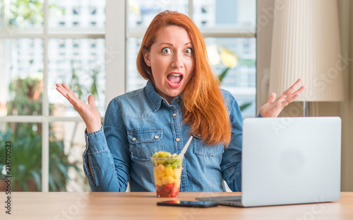 Redhead woman using computer laptop eating fruit at home very happy and excited, winner expression celebrating victory screaming with big smile and raised hands