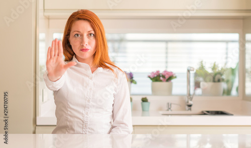 Redhead woman at kitchen doing stop sing with palm of the hand. Warning expression with negative and serious gesture on the face.