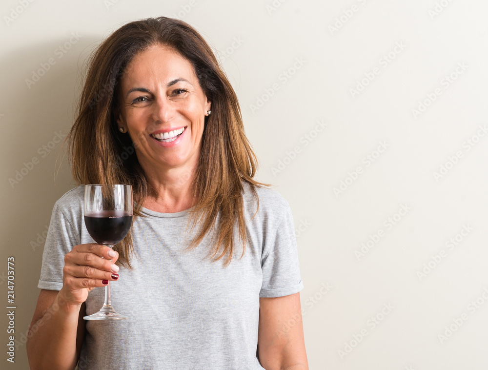 Middle age woman drinking red wine in a glass with a happy face standing and smiling with a confident smile showing teeth Middle age woman drinking red wine in a glass with a happy face standing and smiling with a confident smile showing teeth