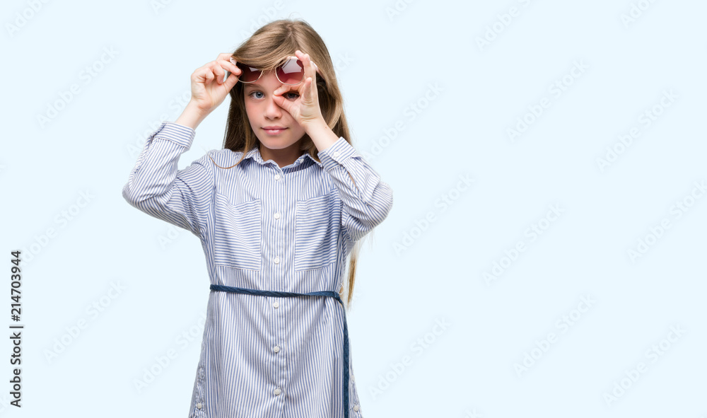 Young blonde toddler wearing blue shirt with happy face smiling doing ok sign with hand on eye looking through fingers Young blonde toddler wearing blue shirt with happy face smiling doing ok sign with hand on eye looking through fingers