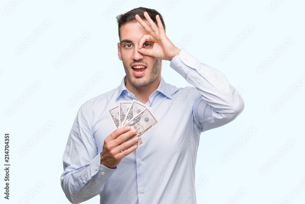 Handsome young man holding money with happy face smiling doing ok sign with hand on eye looking through fingers