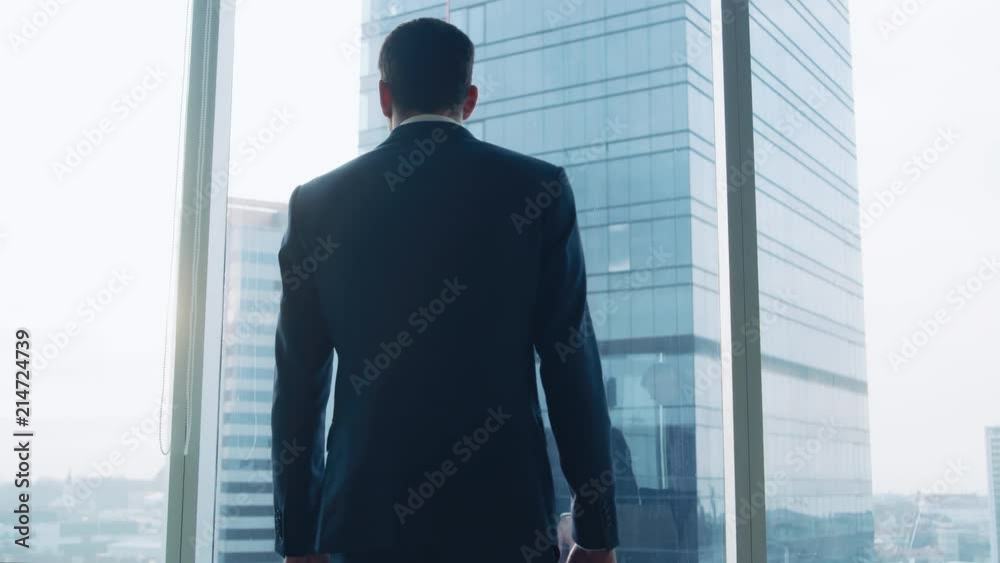 Following Low Angle Elevating Shot of the Confident Businessman in a Suit Walking Through His Office, Putting Hands in Pockets and Looking out of Window. Following Low Angle Elevating Shot of the Confident Businessman in a Suit Walking Through His Office, Putting Hands in Pockets and Looking out of Window.