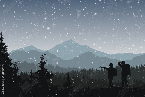 Two tourist on a hike through the winter forest with snowfall and mountains in the background