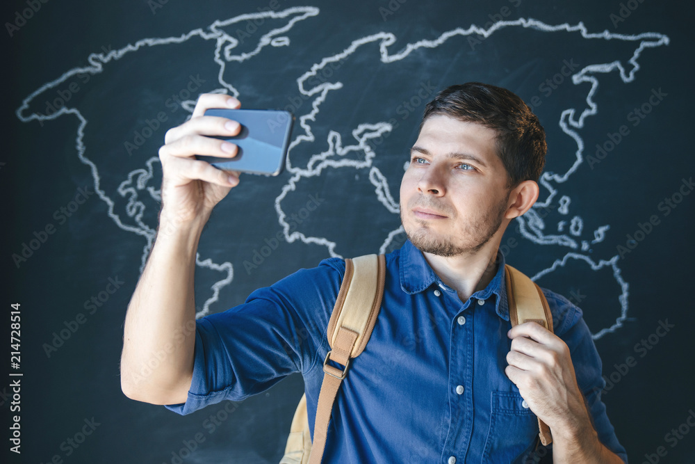 A man in a blue shirt against a background of a dark chalky wall with a smartphone in his hand and a backpack, makes a photo.